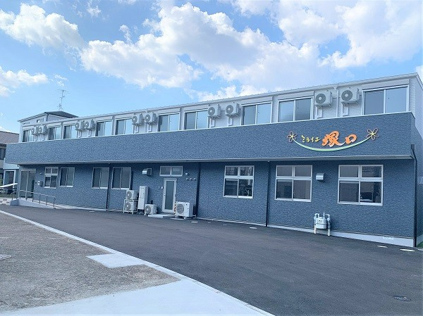 白とダークグレーの２階建ての建物は横に長く、玄関先には手すり付きのスロープと階段がある。