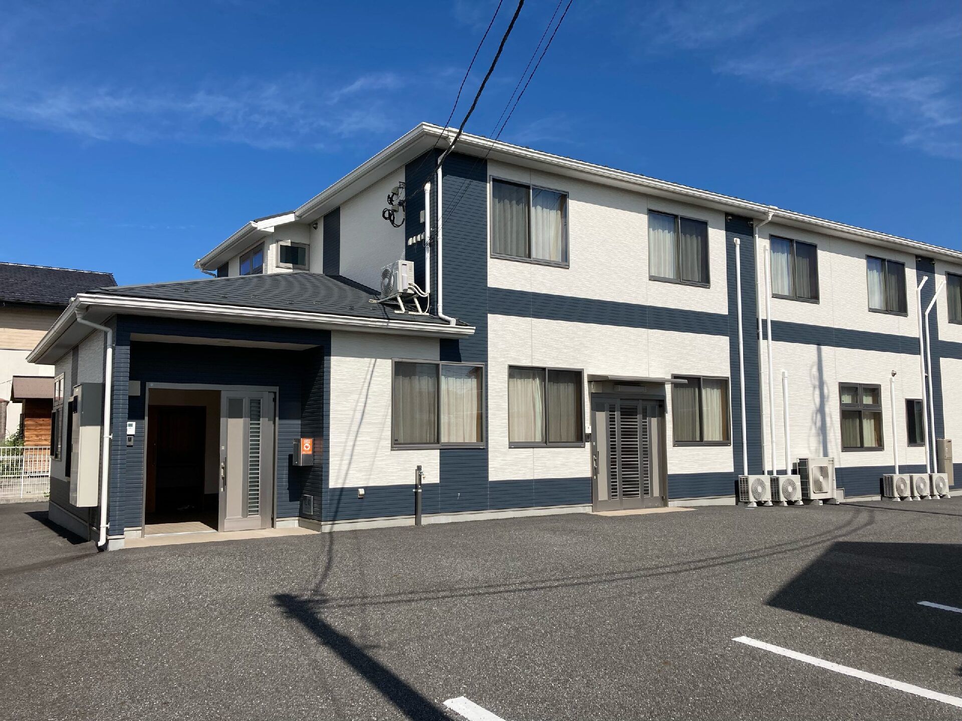 広い駐車場がある敷地内にはダークブルーと白の２階建ての建物が建っている。玄関には屋根も付いている。