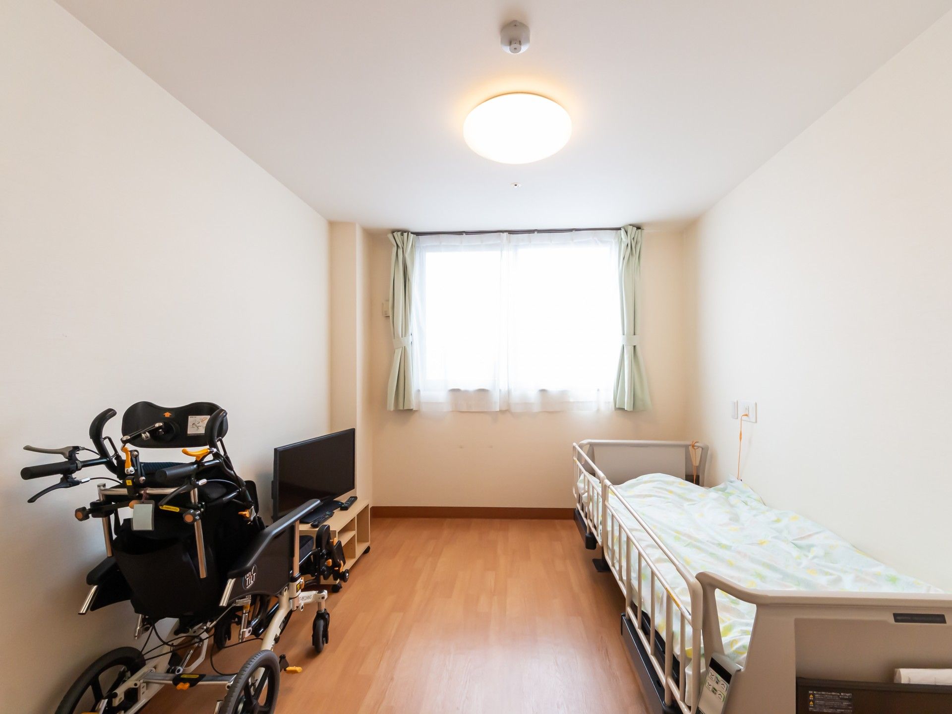 レースカーテンとカーテンが付いた窓があるフローリングの部屋には寝具付きのベッドやシルバーカーなどが付いている。