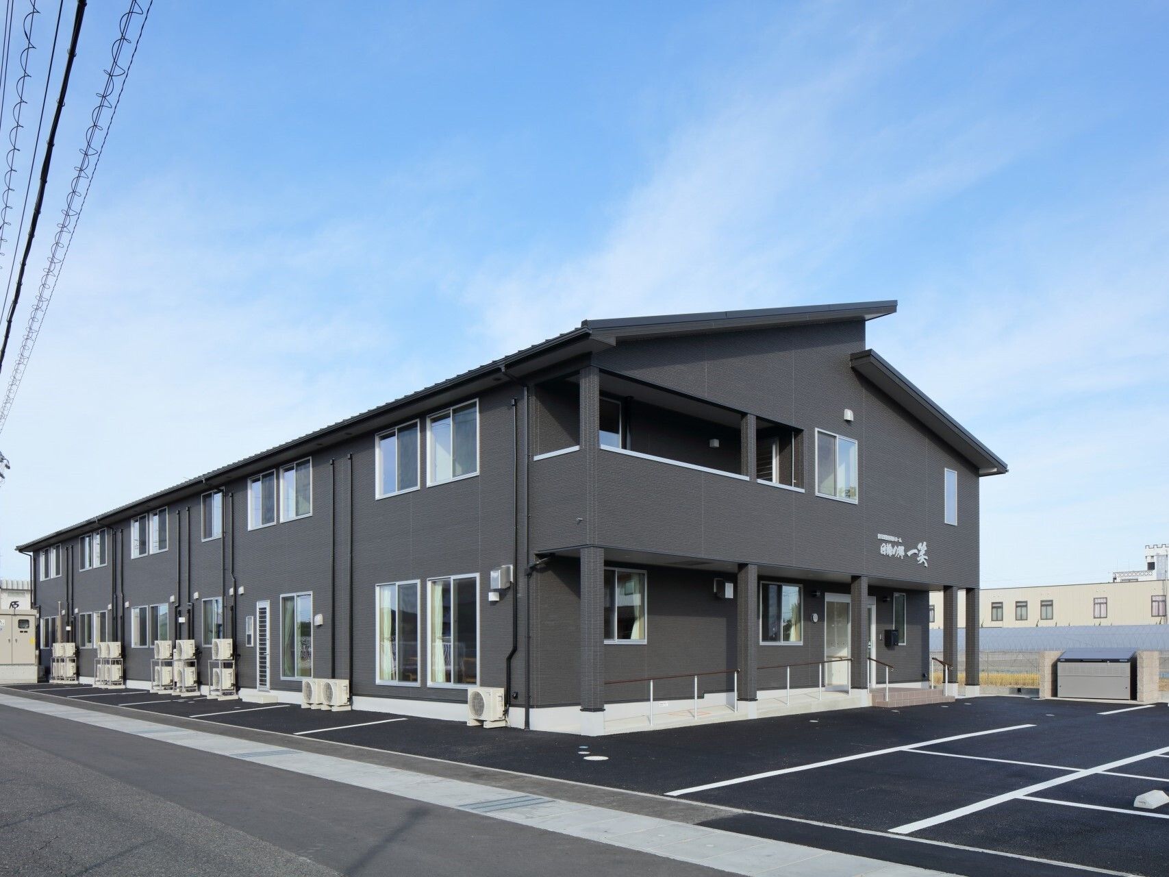 ダークグレーの外壁の2階建ての建物。敷地内には駐車場もあり、日当たりのいい場所にある。
