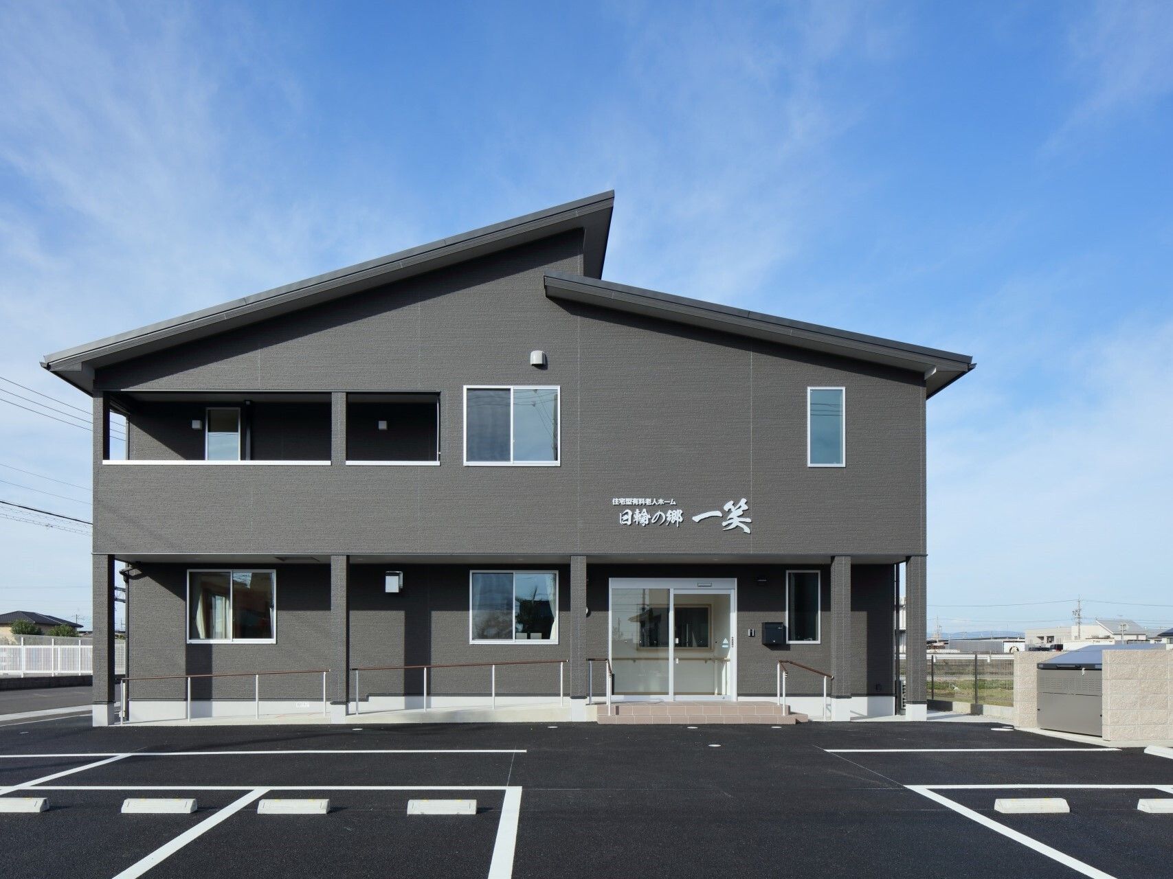広い駐車場が付いたダークグレーの２階建ての建物は日当たり良好。玄関先には手すり付きのスロープもある。