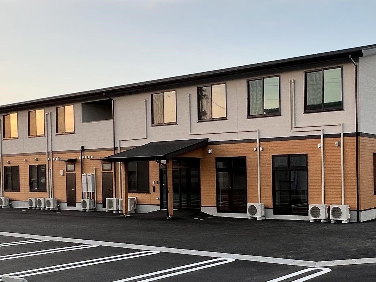 2階部分はベージュ、1階部分はブラウンの２階建ての建物は日当たり良好。玄関には屋根が付いている。