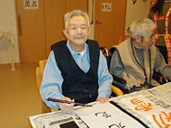 高齢男性がいすに座り、筆を持って習字をしている様子。テーブルには半紙が置かれ、新聞が敷かれている。