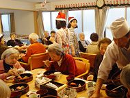 食卓を囲んでいる入居者たちの様子。サンタ帽をかぶった女性スタッフが数名食堂を歩いている。