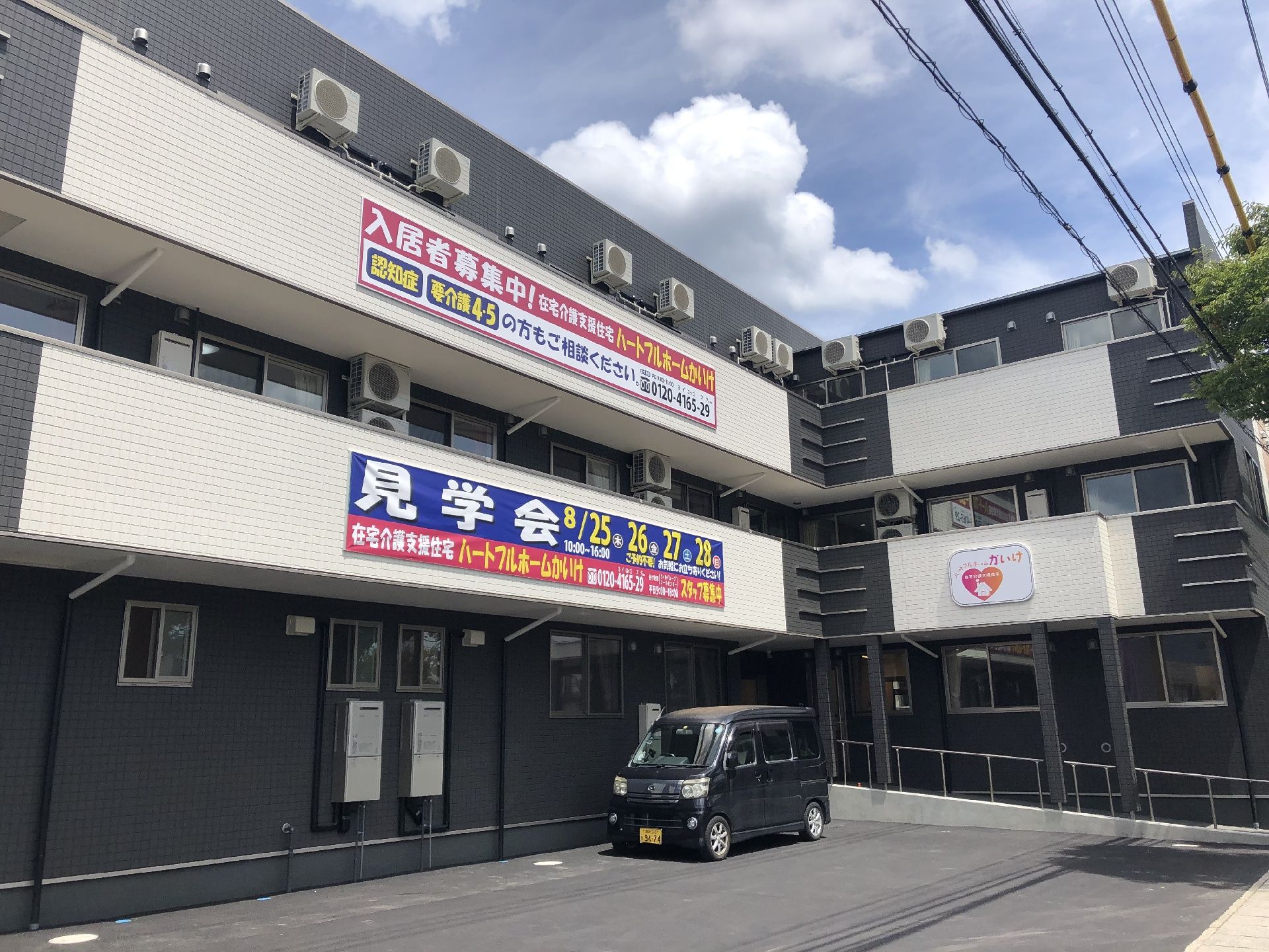 ダークグレーの3階建ての建物はエル字型で日当たりのいい場所にあり、敷地内には車が止まっている。
