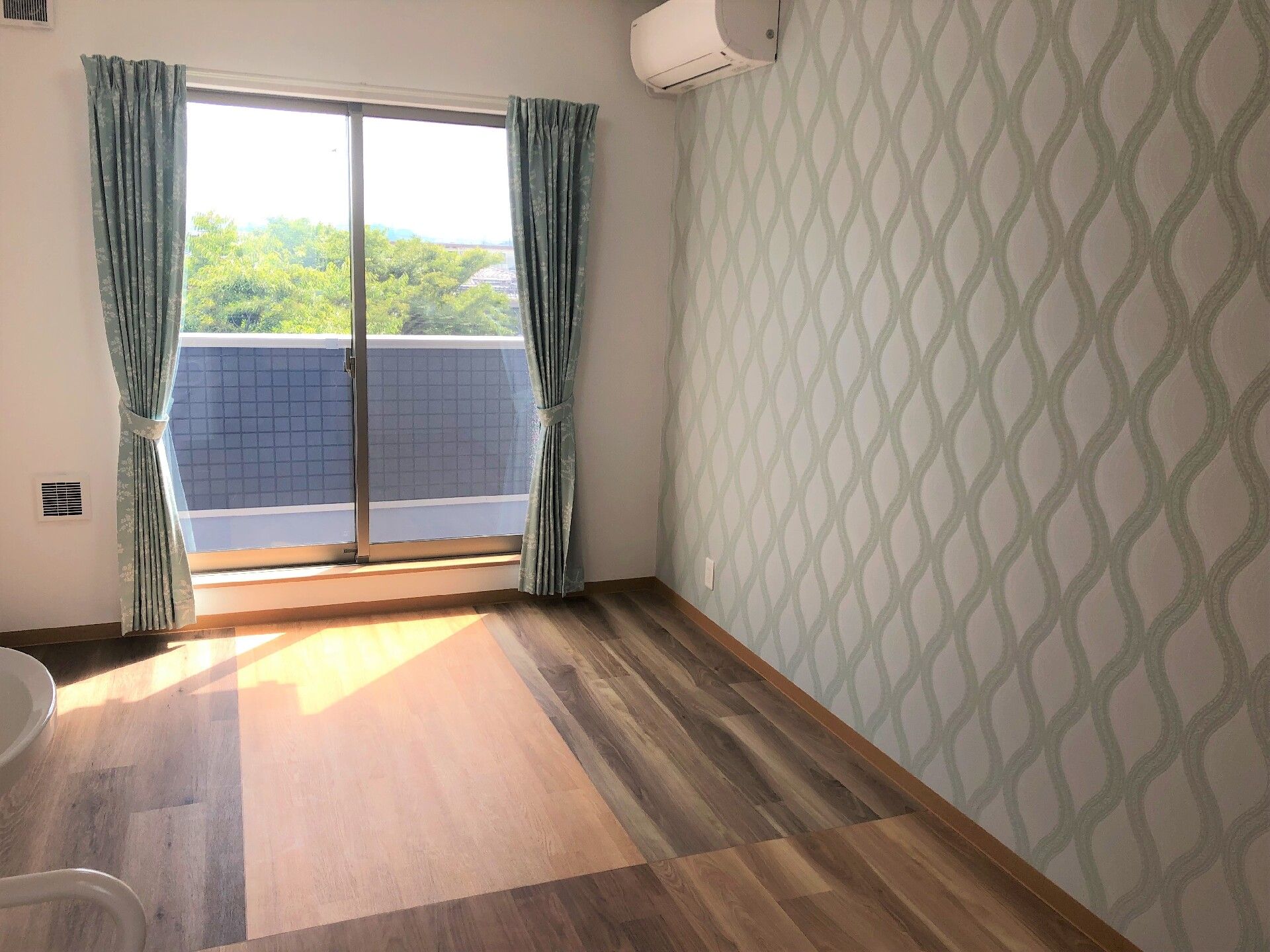 ベランダが付いたフローリングの部屋にはカーテンがある窓があり、エアコンも付いている。