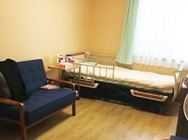 介護ベッドが置かれたフローリングの部屋。一人用の椅子が設置され、カーテン付きの窓がある。
