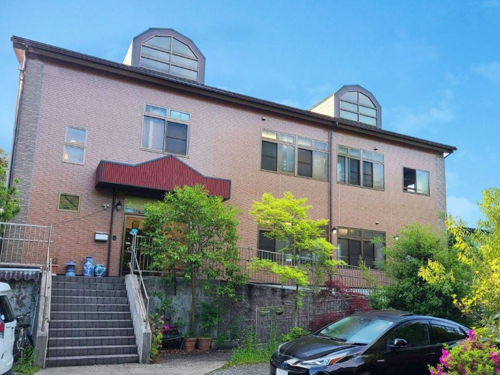ブラウン系の２階建ての建物は日当たり良好で建物の前には階段があり、駐車場も付いている。