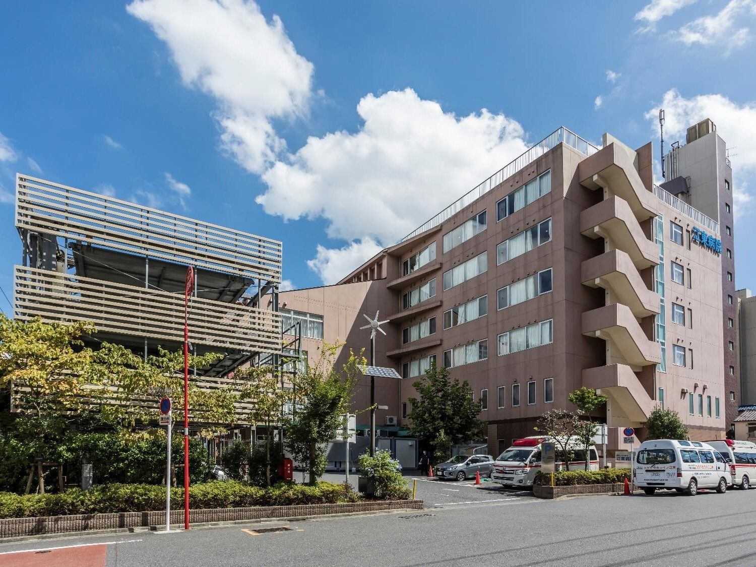 ブラウン系のエル字型の大きな建物が道路に面して建っている。駐輪場には救急車もある。