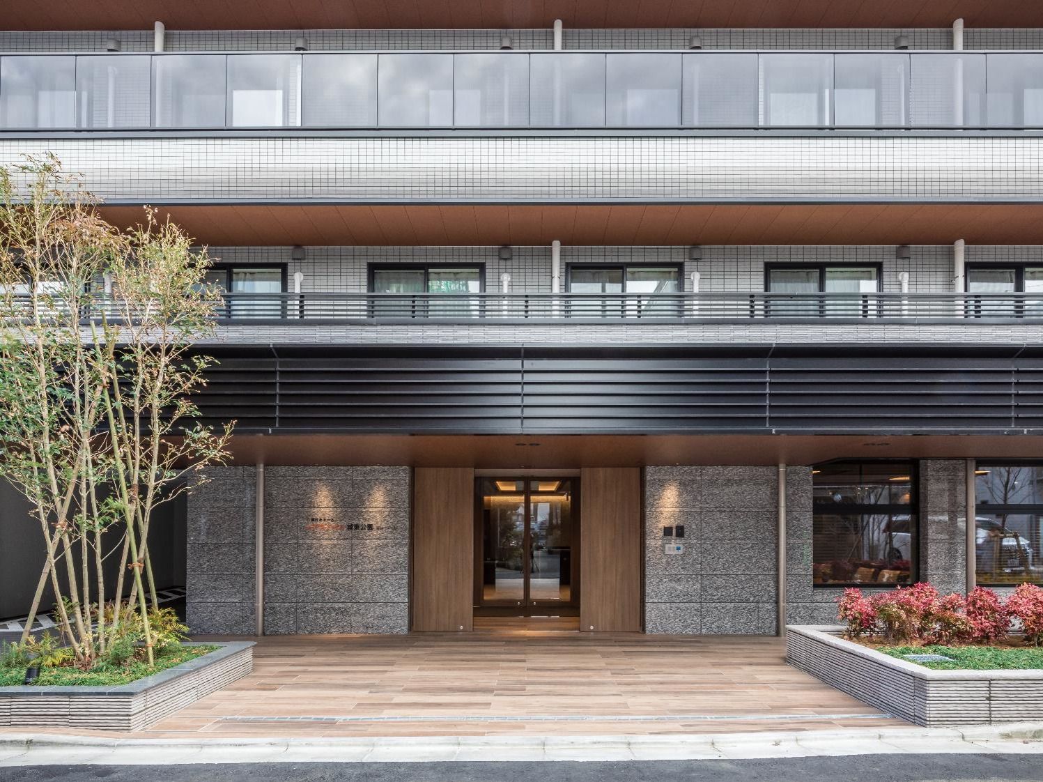 各フロアにベランダがあり、グレー系の外観のしっかりとした建物。玄関周りには草木が植えられている。