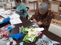 真剣な表情で創作活動をしている高齢女性たち。テーブルの上には色紙などが置かれている。