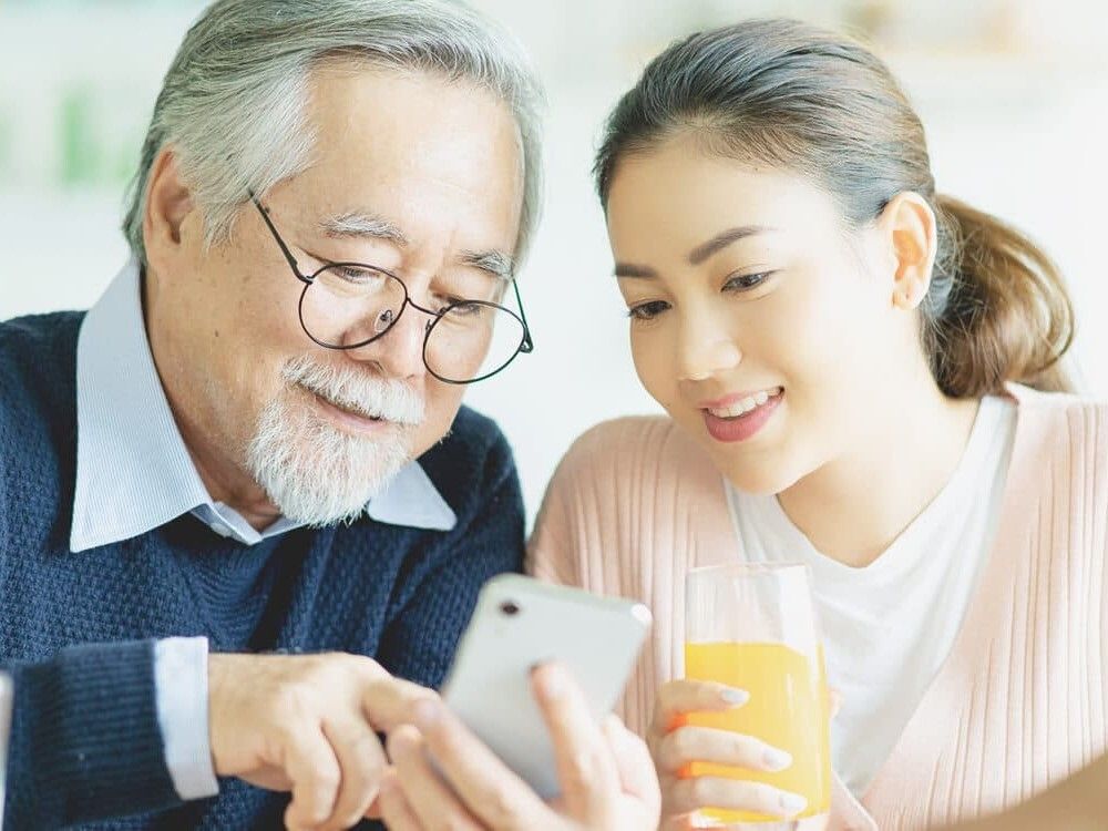 オレンジジュースを持った若い女性と笑顔でスマホを見ながら話している眼鏡をかけた高齢男性。