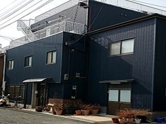 ダークブルーの外壁の建物の周りには草木が植えられた鉢がたくさん置かれ、日当たりのいい場所にある。