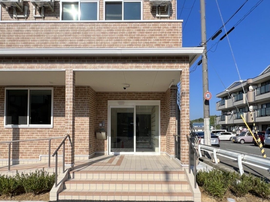レンガ調の２階建ての建物は横に長く、玄関には緩やかなスロープと階段があり、手すりも付いている。