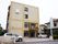 施設の建物は4階建てで、玄関前には屋根がせり出している。外壁はイエロー系の濃淡で、左側上部にはロゴマークが表示されている。