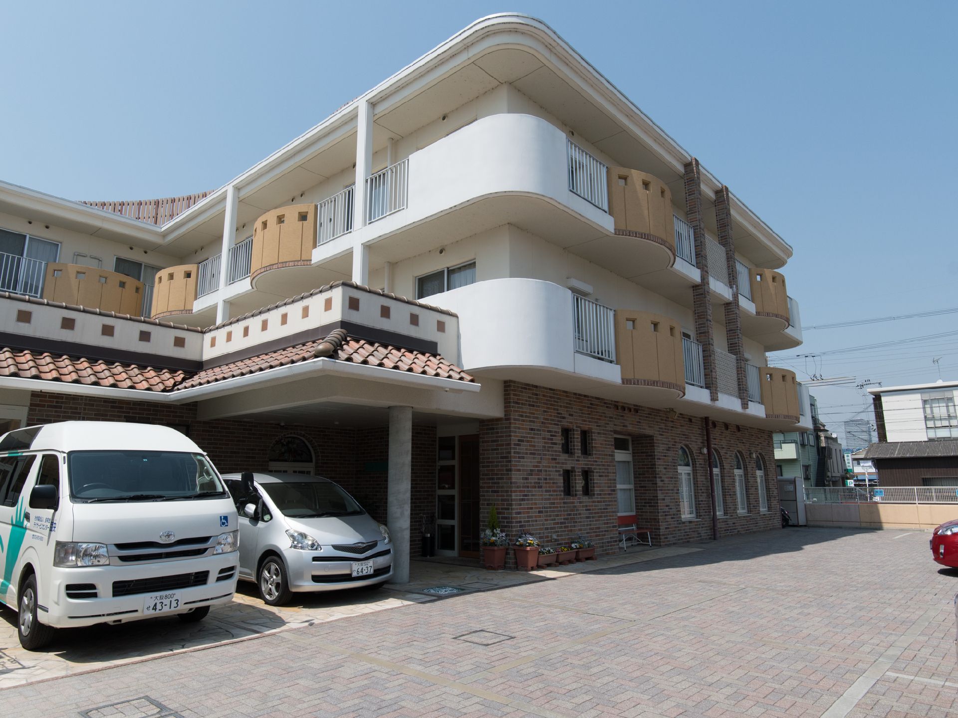 正面に3階建ての建物がある。左側には2台の車が止まっている。右側は駐車場になっている。玄関には外屋根が付いている。