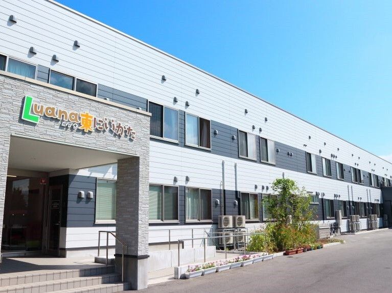 車寄せや手すり付きのスロープ、階段が付いた２階建ての建物は日当たり良好でグレー系の外観。