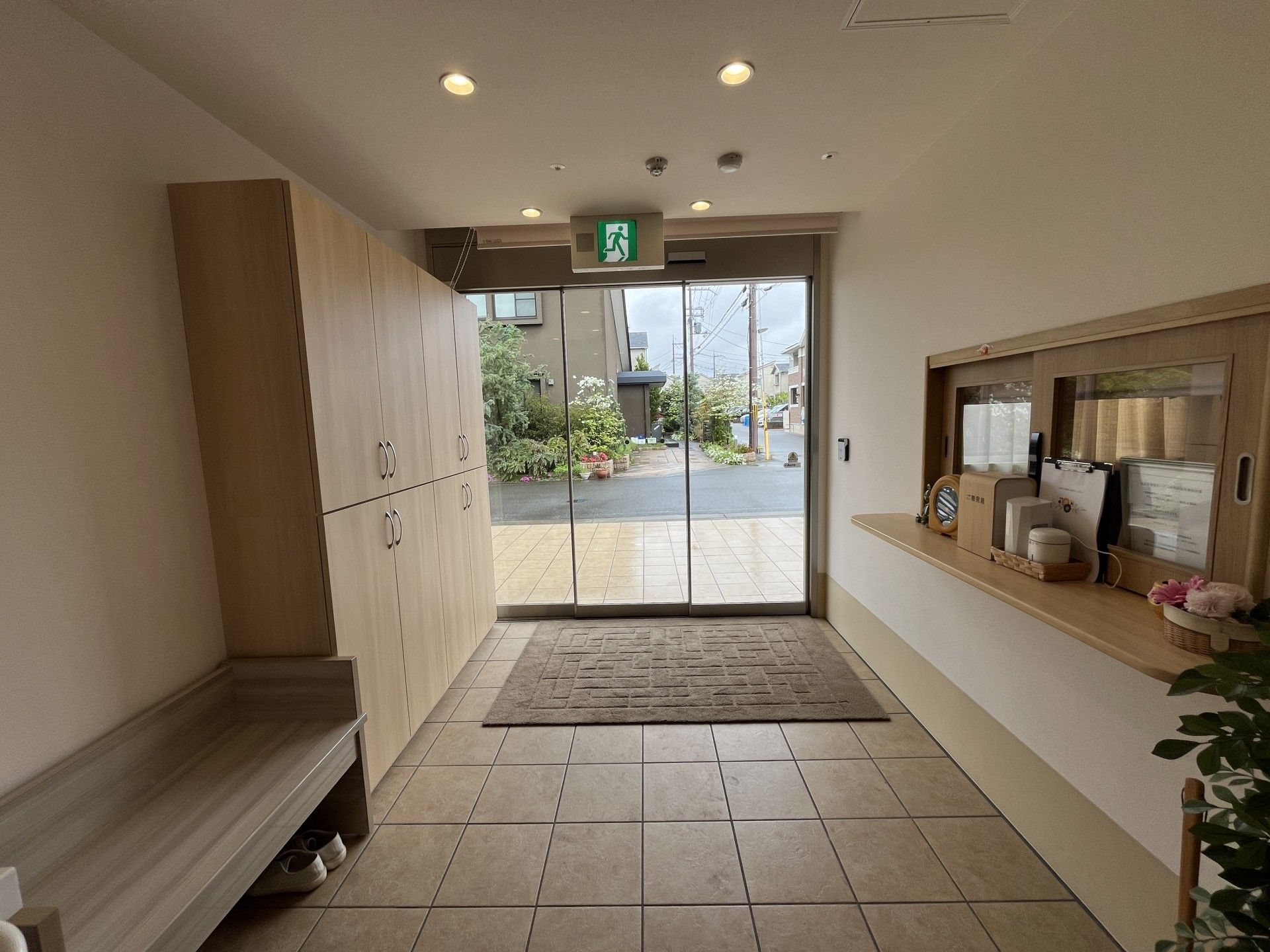 グレーの屋根が付いたブラウン系の外壁の２階建ての建物は日当たりがいい場所にあり、玄関先には手すり付きのスロープと階段がある。