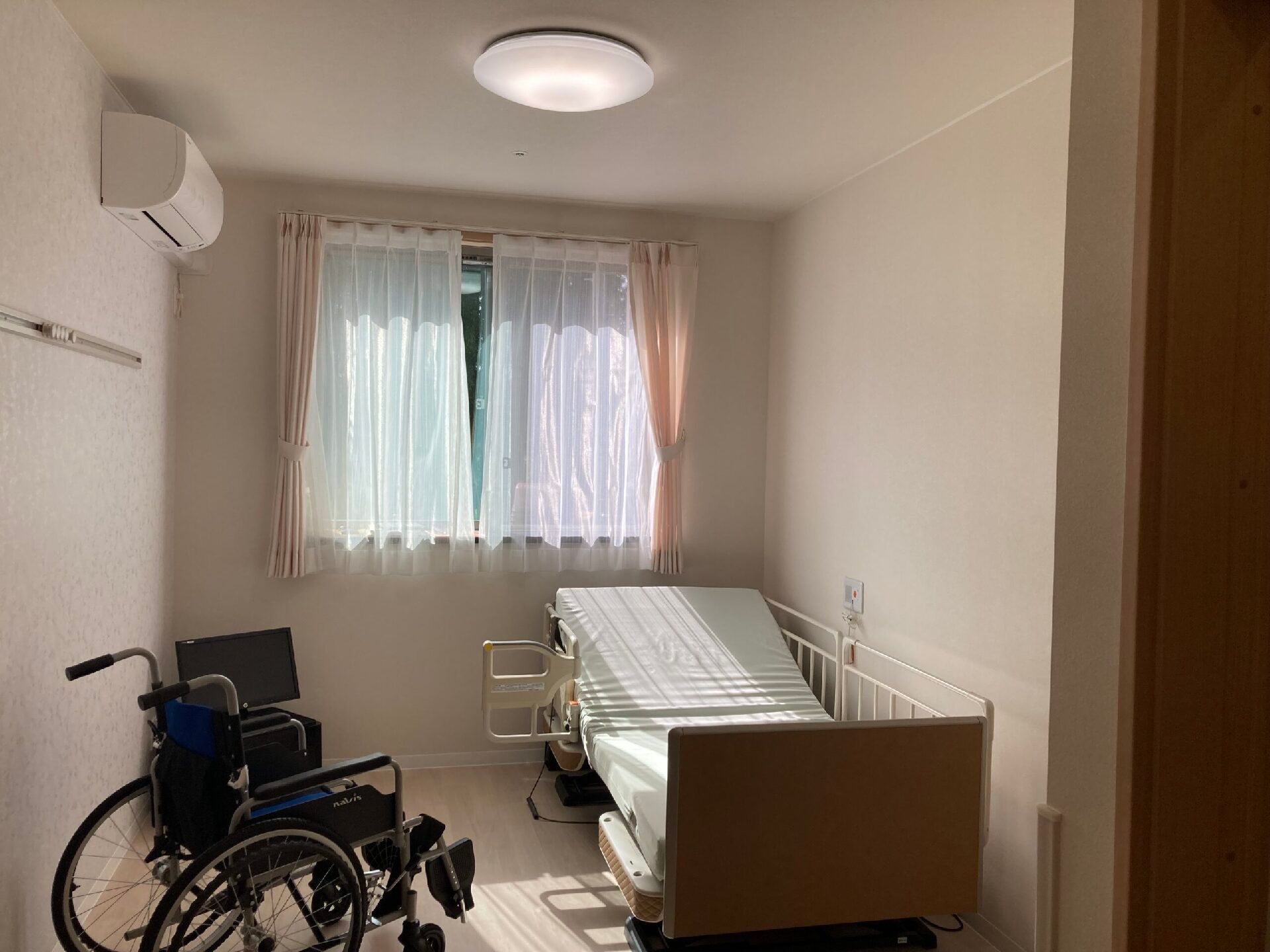 介護ベッドや車いすが置かれたフローリングの部屋にはカーテン付きの窓があり、緊急通報装置もある。