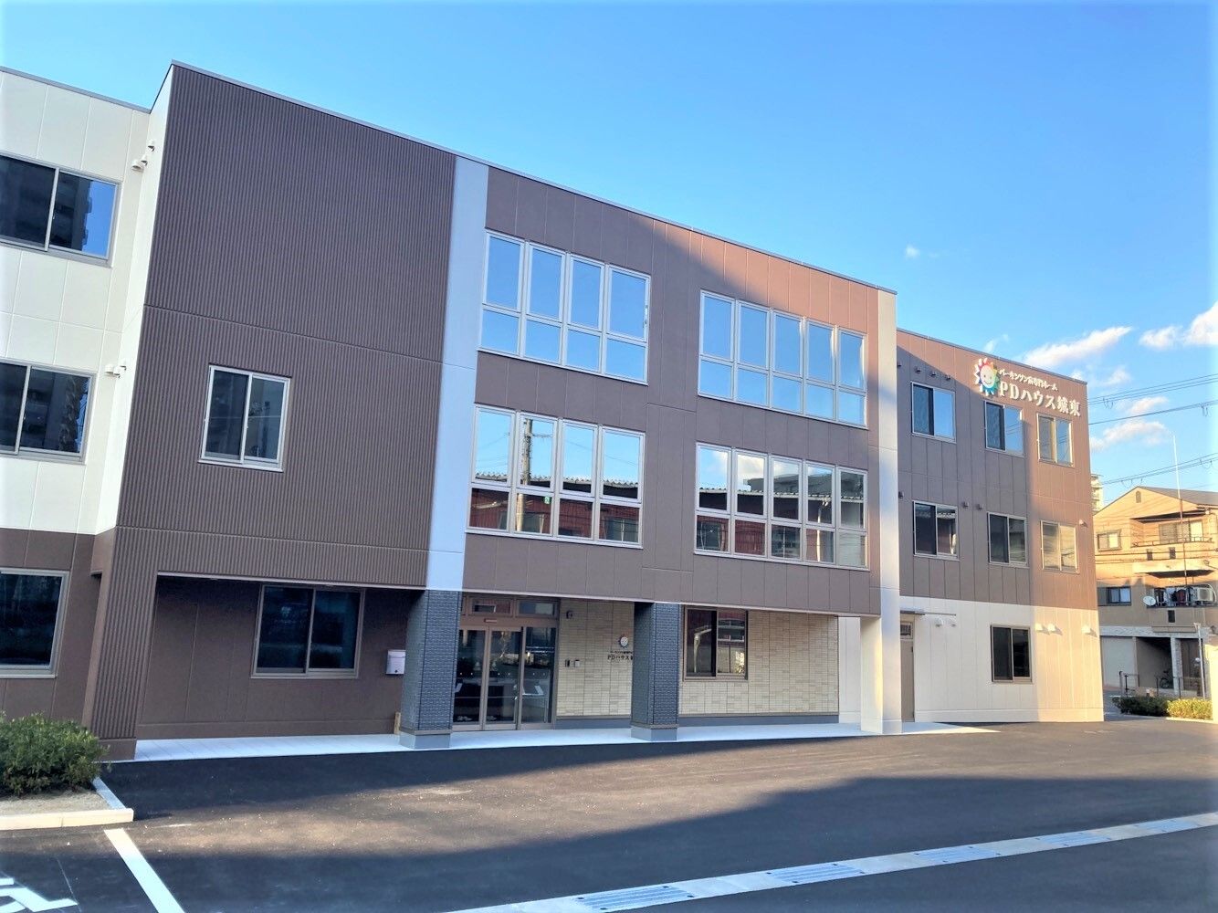 ベージュとブラウンの外壁の3階建ての建物。日当たりのいい場所にあって、周りには草木も植えられている。