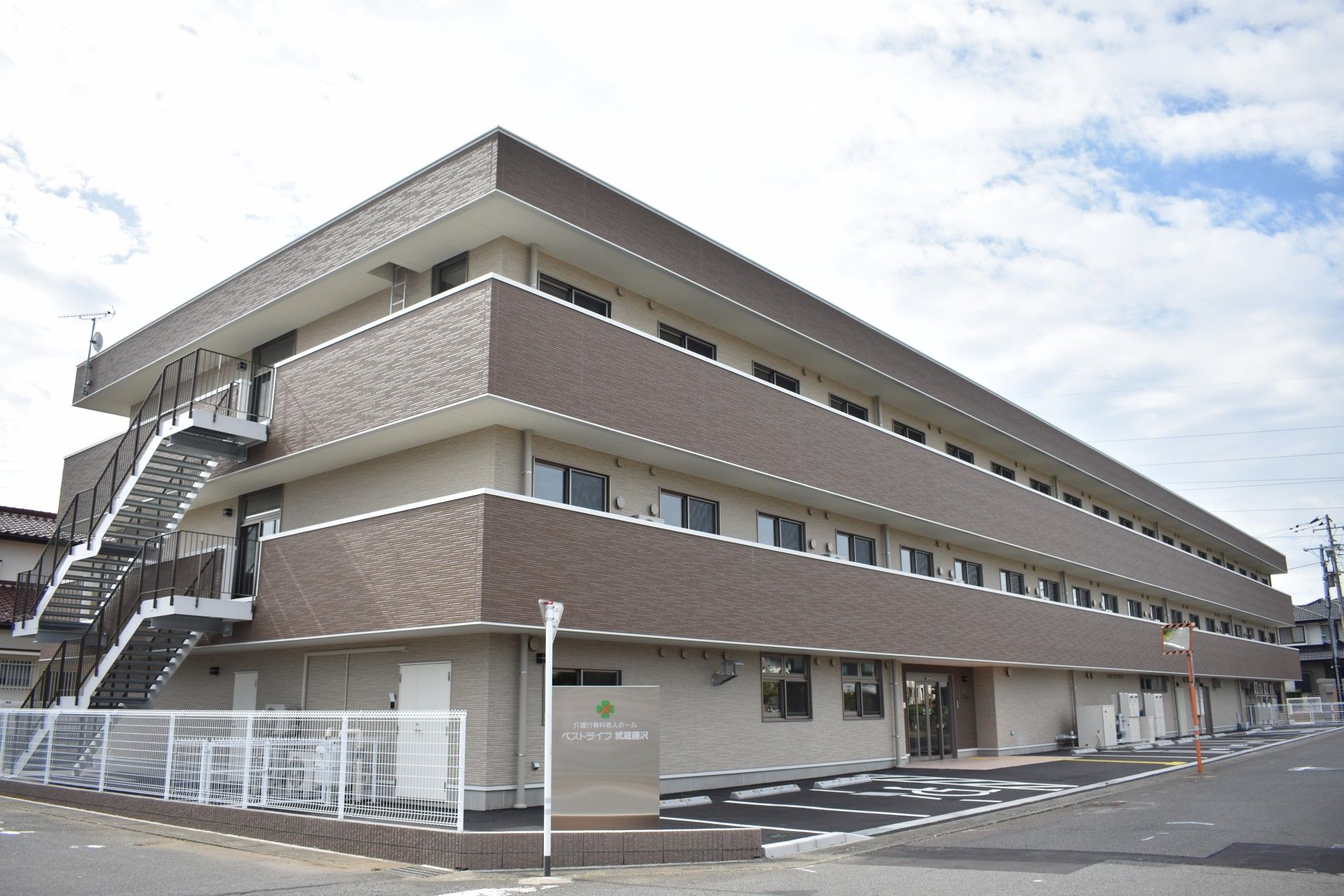 駐車場が付いた3階建ての建物はブラウン系で屋外階段やベランダが付き、日当たり良好。