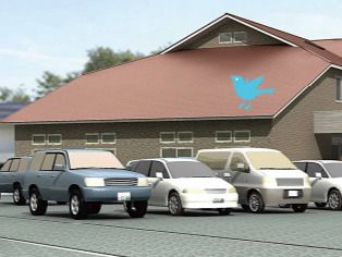 車がたくさん止まっている駐車場があり、その奥にはブルーの鳥の絵が書かれた屋根が付いたブラウン系の建物がある。