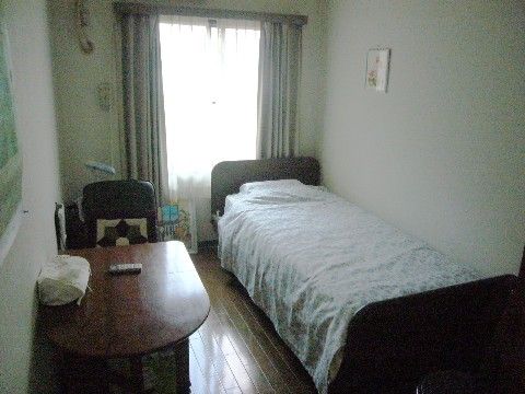 窓から外光が入る、落ち着いた部屋。エアコン付きで、木製の低床型のベッドと一人用のダイニングセットがある。