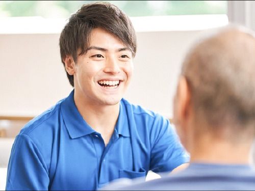 手前には高齢男性の後ろ姿があり、その向こうには笑顔で話しているブルーのユニフォームを着た男性スタッフがいる。
