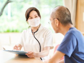 眼鏡をかけた高齢男性のそばにはカルテのようなものを見ながら話しているマスクをした女性スタッフがいる。