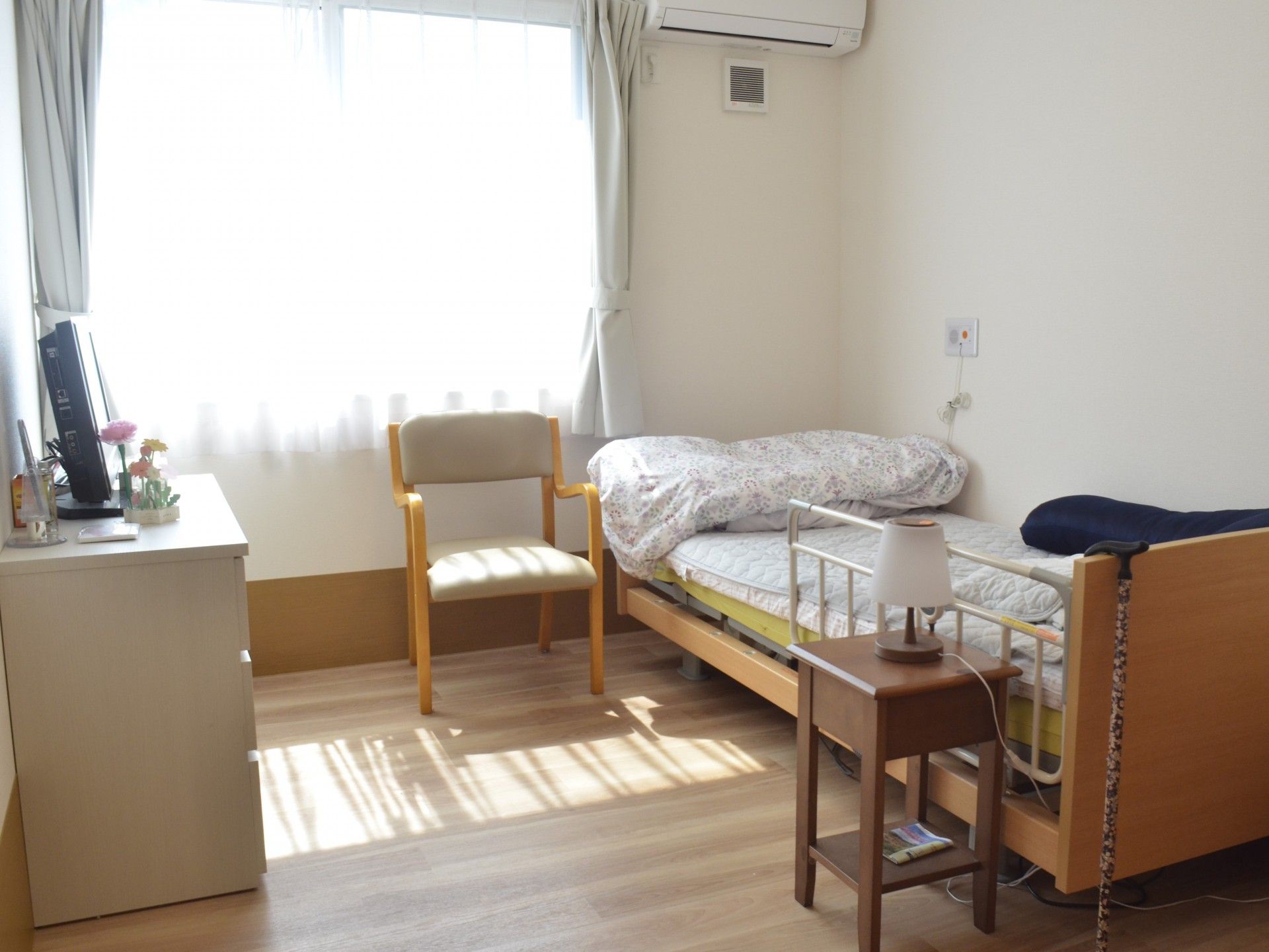 レースカーテンとカーテンが付いた大きな窓があるフローリングの部屋には介護ベッドやエアコンがあり、タンスも置かれている。