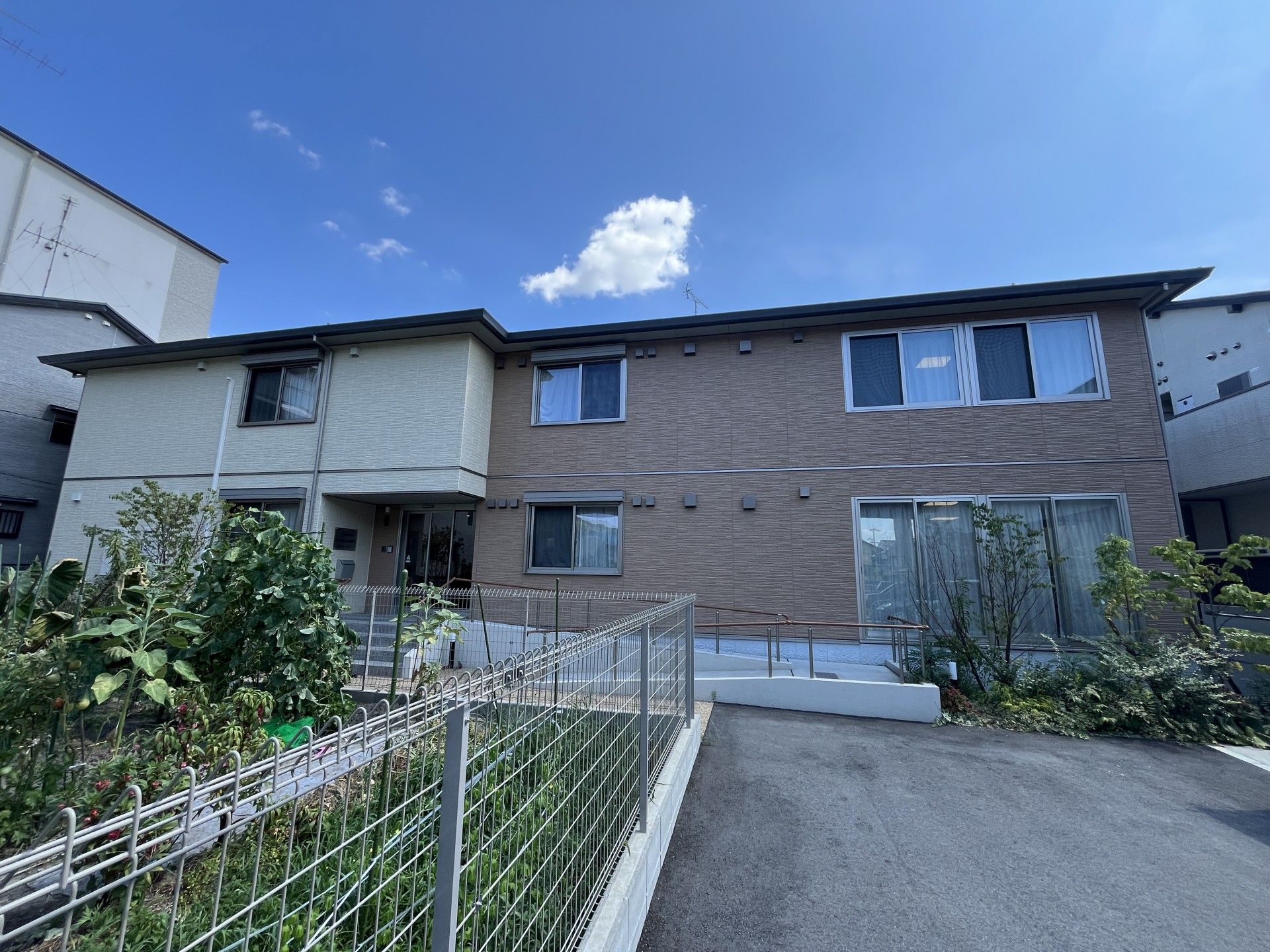 駐車場がある２階建ての建物は横に長く、ベージュとブラウンの外壁。玄関先には手すり付きのスロープと階段がある。