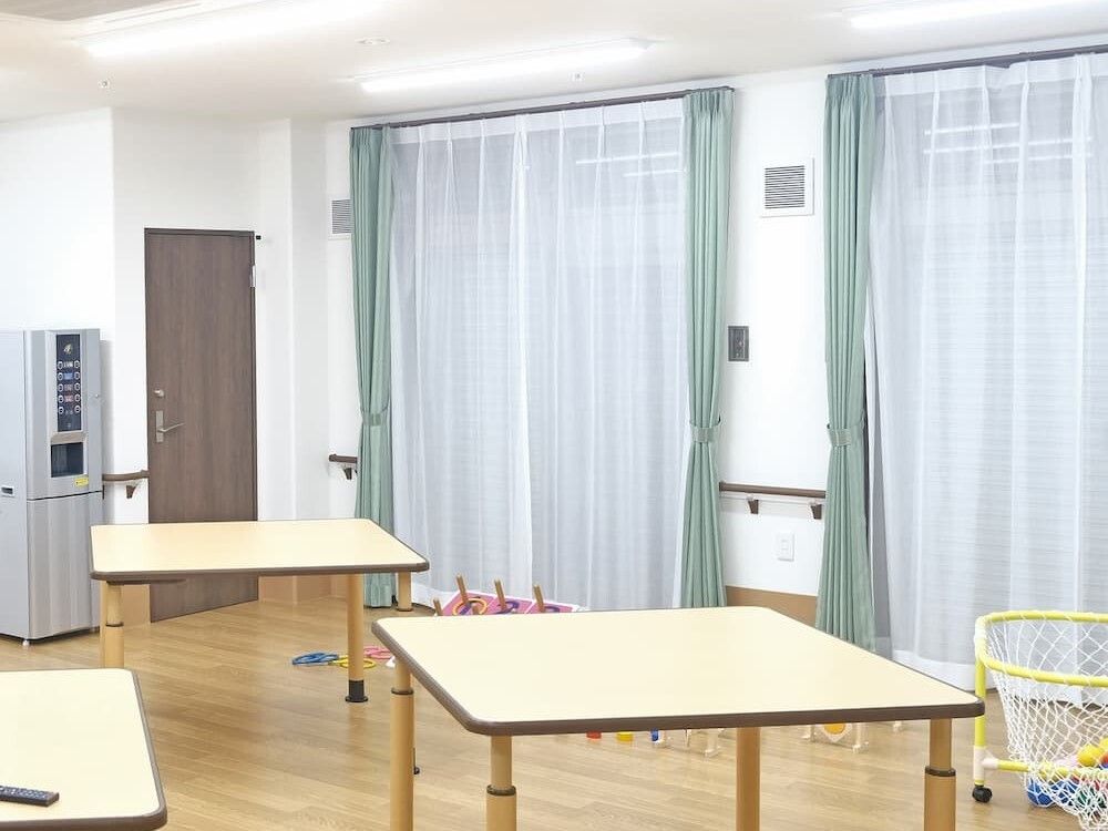 レースカーテンとカーテンが付いた大きな窓があるフローリングの部屋にはテーブルが複数あり、輪投げなどもある。