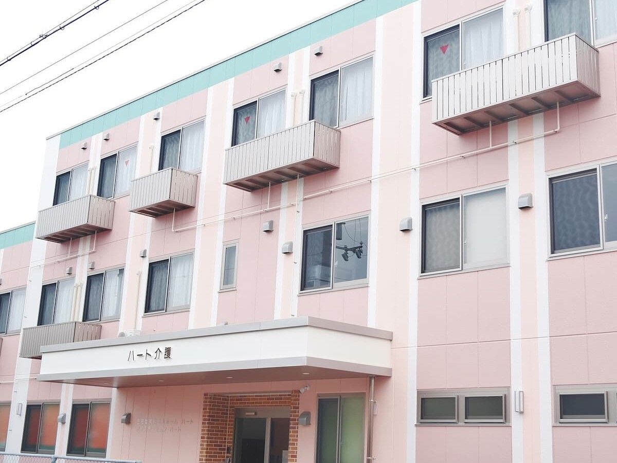ピンクの外壁の３階建ての建物は玄関先にホーム名が書かれた屋根が設置されている。