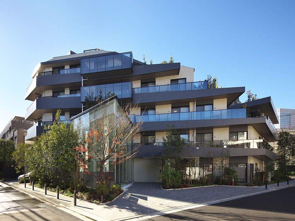 角フロアにベランダが付いたダークグレーの外壁の建物は大きく、建物の周りには草木が植えられている。