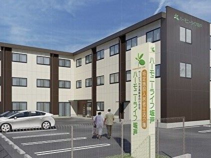 3階建ての建物はエル字型で窓がたくさんあり、白とダークグレーの外壁。駐車場内を高齢夫婦が歩いている。