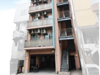 ブラウン系の外壁の建物は縦に長く、右側に階段があり、左側にベランダ付きのフロアがある。