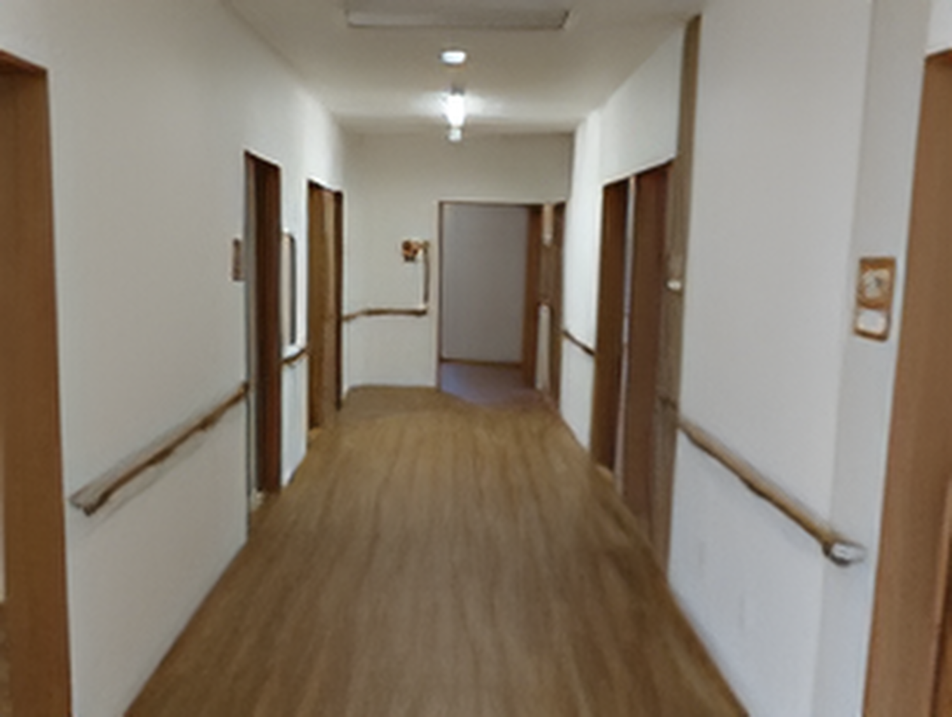 まっすぐな廊下の両サイドに手すりと居室のドアがあり、天井には照明が点いている。
