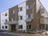 白とブラウンの3階建ての建物は角フロアにベランダが付いていて、玄関には屋根があり、建物の周りには草木が植えられている。