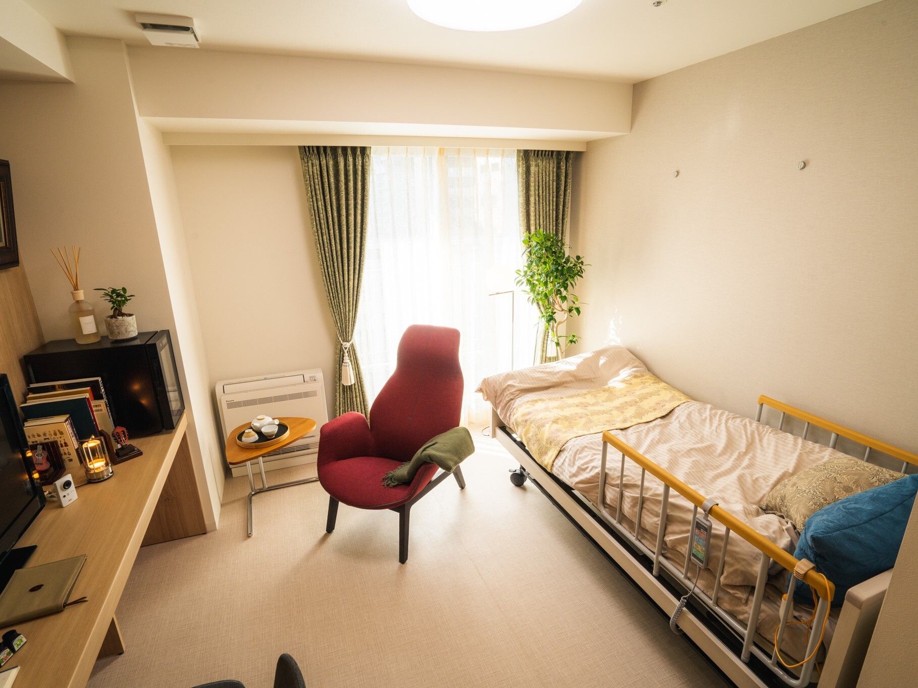 寝具付きのベッドが置かれ、レースカーテンとカーテンが付いた大きな窓が付いている部屋にはいすやテーブルもある。