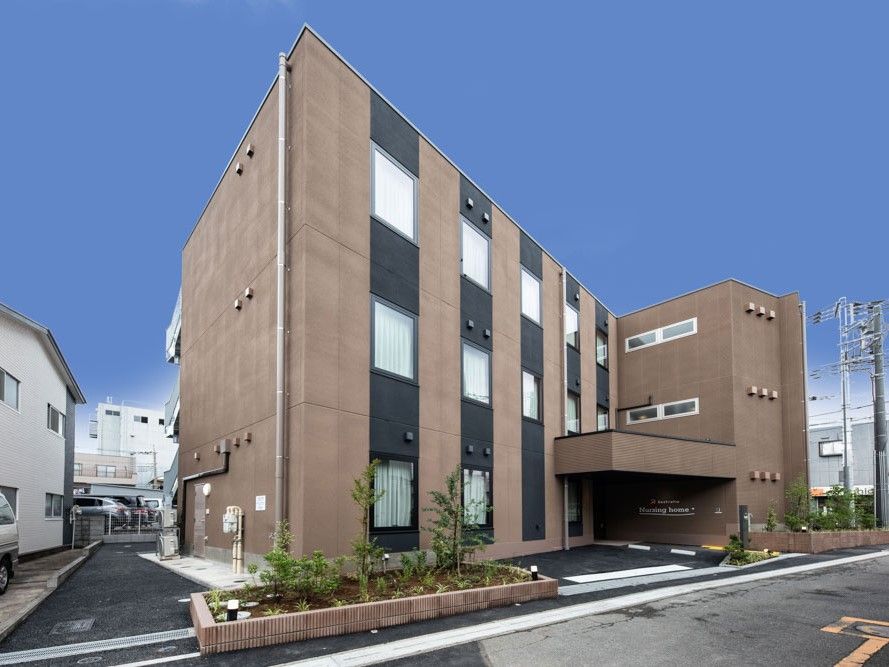 ブラウン系の外壁の3階建ての建物の玄関先には屋根があり、外壁にはホーム名が書かれている。