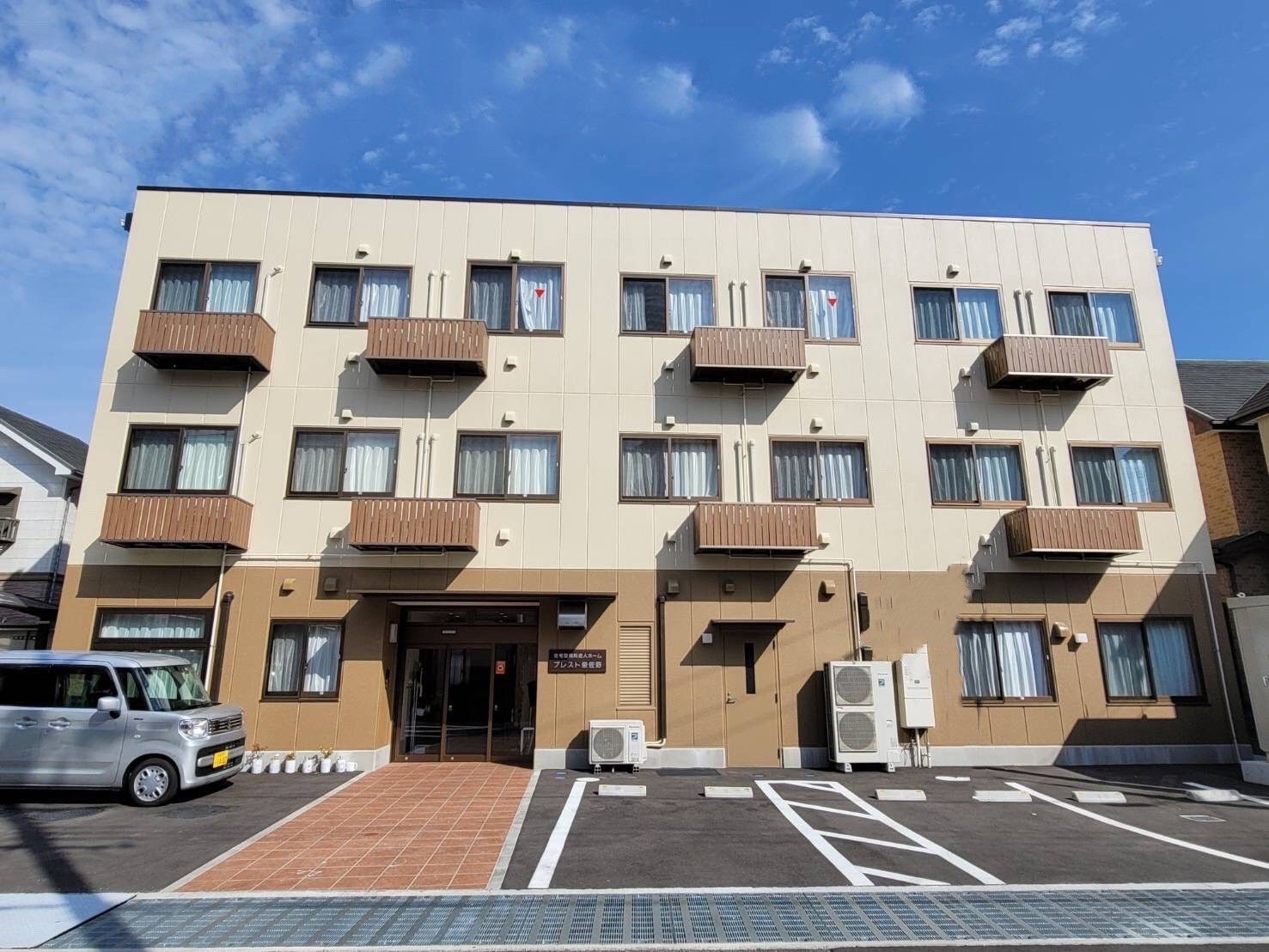 白とブラウンの3階建ての建物は日当たりのいい場所に建っていて、建物の前には駐車場もある。