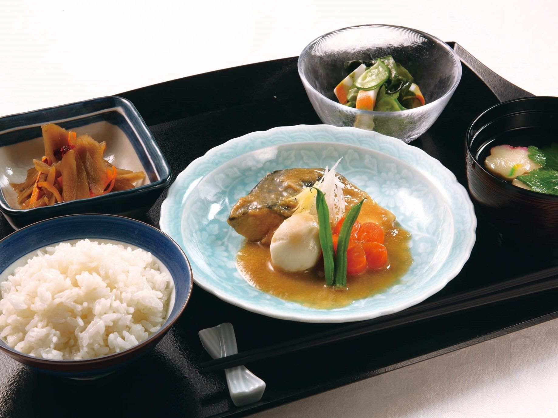 黒いトレーには煮物付きの魚の煮付けや野菜の和え物、吸い物、白いご飯などが並んでいる。