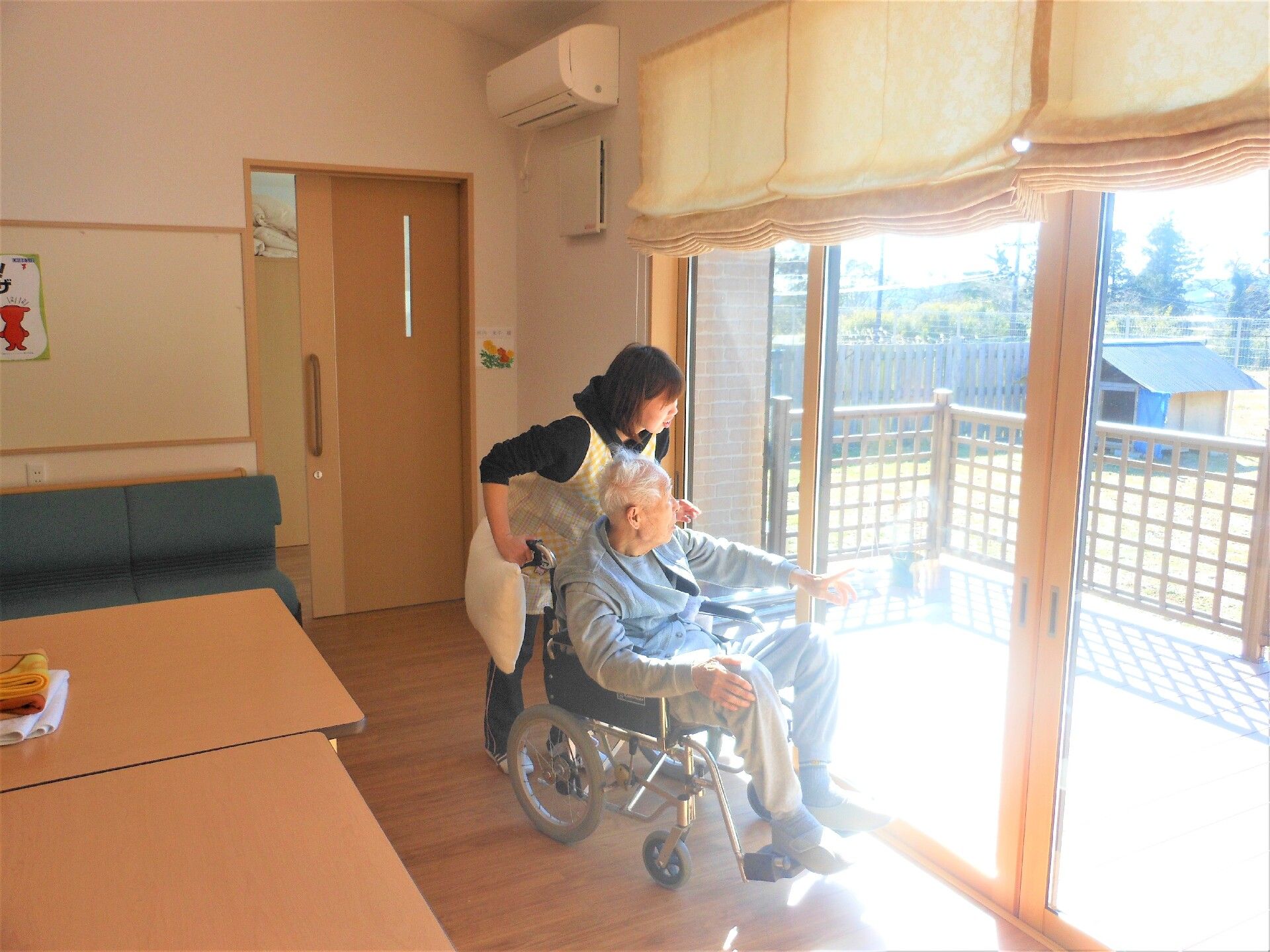 車いすの高齢男性と付き添いのエプロンを付けた女性スタッフが室内の窓から外にあるテラスの方を見ている。