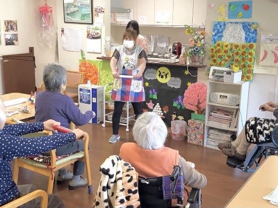 椅子に座って帽を持っている高齢女性たちが前にいる女性スタッフを見習ってエクササイズをしている。