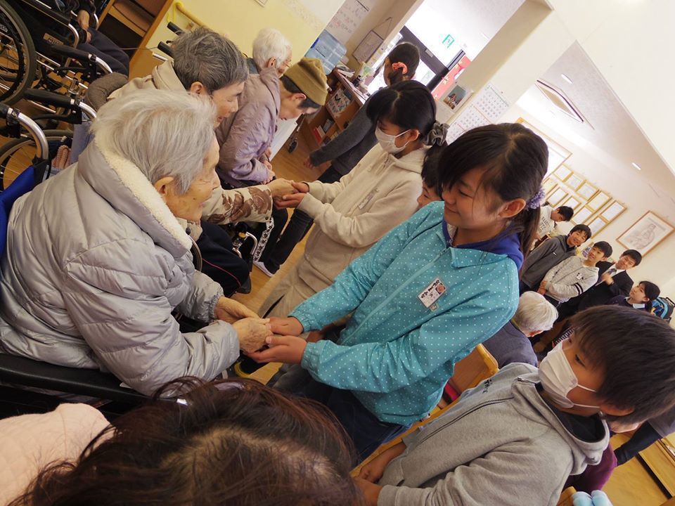 名札を付けた小学生たちと手を取り合って笑っている車いすの高齢女性たち。