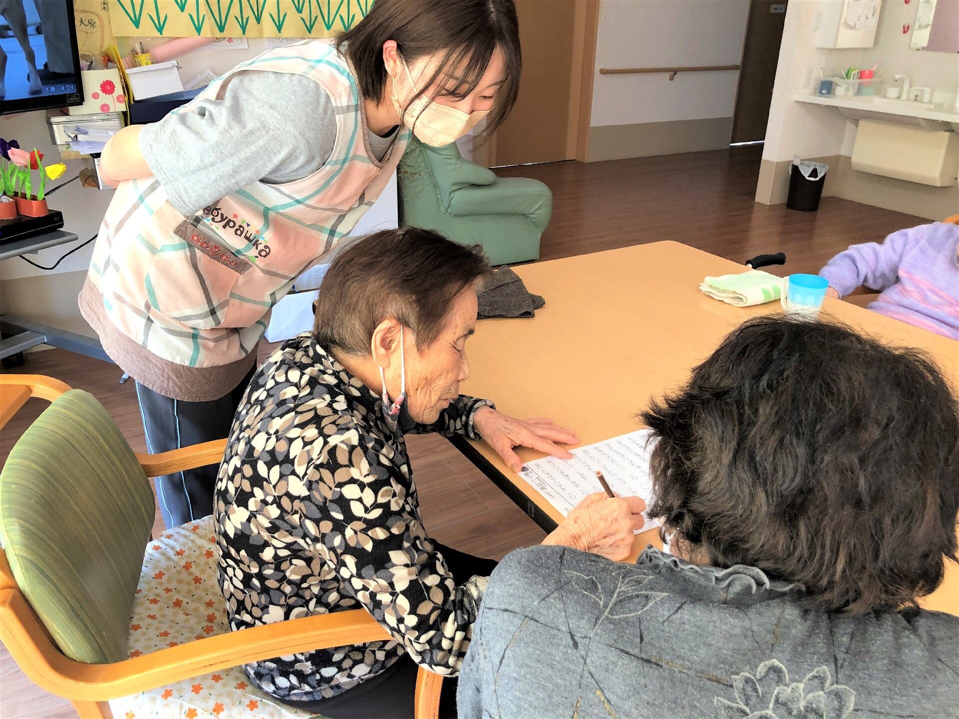 紙になにかを書いている高齢者の両サイドには若いスタッフや高齢女性がいて、注目している。