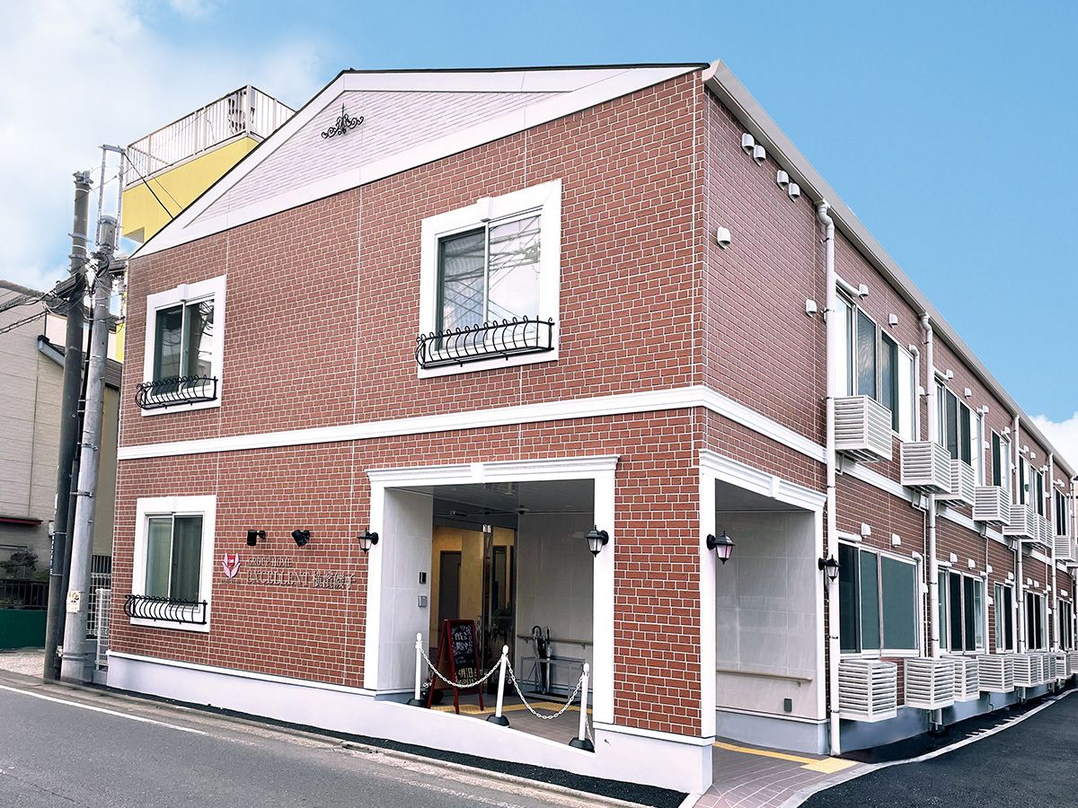 ブラウン系の外観の２階建ての建物の前には一人の人が歩いている。三角屋根が付いた建物は日当たりのいい場所に建っている。