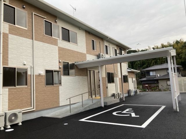 眼鏡をかけた高齢男性が福祉車両から降りようとしていて、エプロンを付けたスタッフが手伝っている。