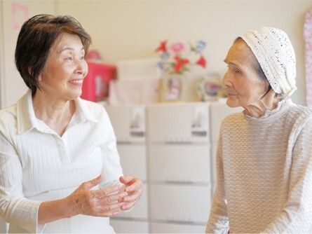 白い家具が置かれているスペースで笑顔で話している白いポロシャツを着た女性スタッフと白い帽子をかぶった高齢女性。