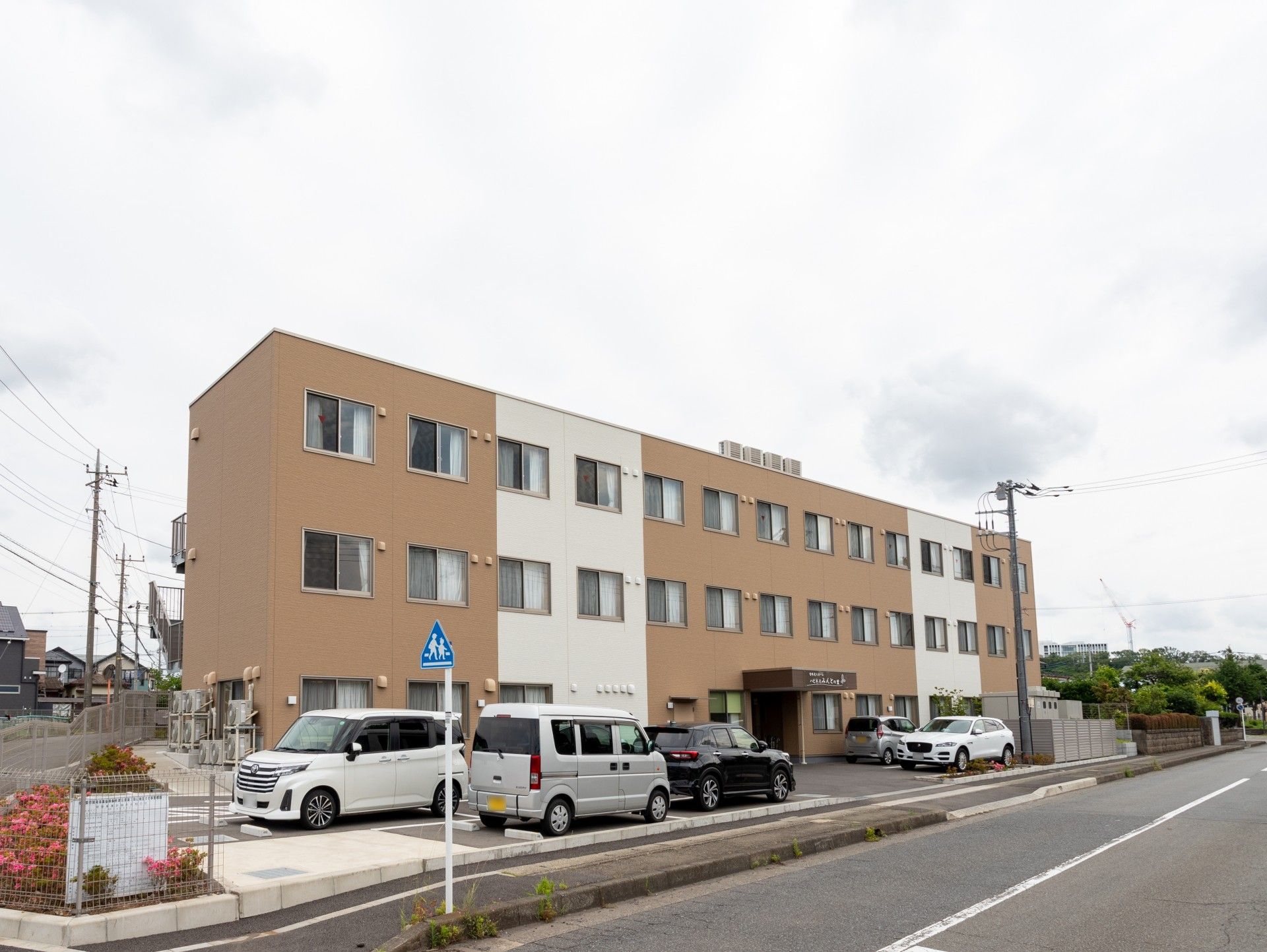 ブラウン系の外観で日当たりのいい場所に建っている3階建ての建物の前には車が止まっている駐車場もある。